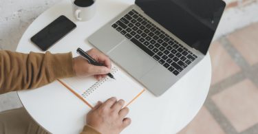 mano de persona escribiendo en una libreta al lado de un ordenador y un teléfono móvil y un café