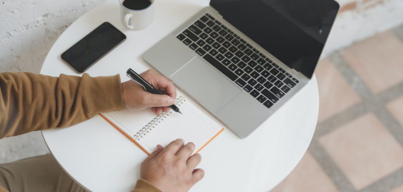 mano de persona escribiendo en una libreta al lado de un ordenador y un teléfono móvil y un café