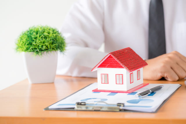 casita encima de papeles con una planta al lado y un hombre con corbata detrás